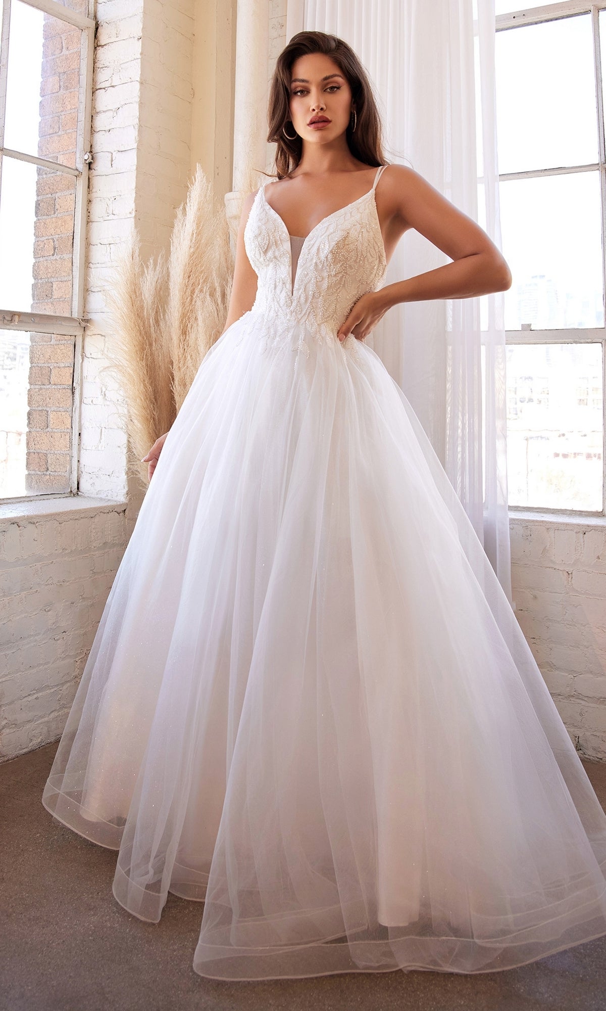 A woman stands indoors by large windows, wearing the Off-White Long A-Line Wedding Dress CD0154W—a sleeveless gown with a fitted bodice and full tulle skirt.